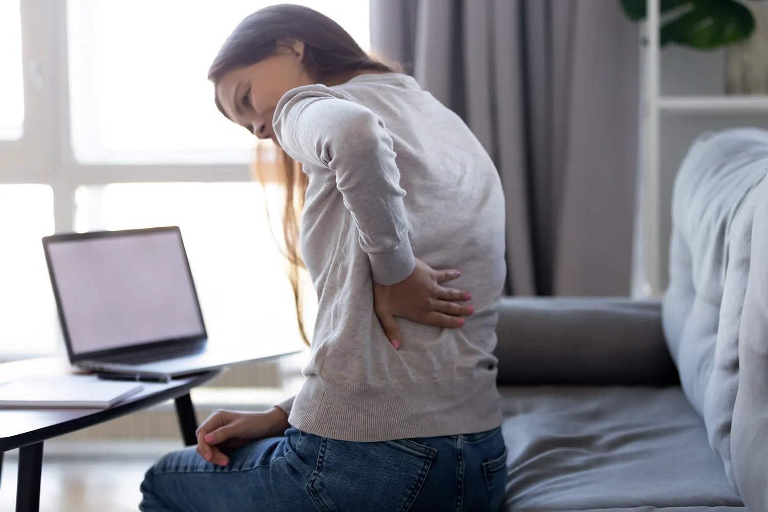 Woman sitting on a couch holding her lower back beside a laptop